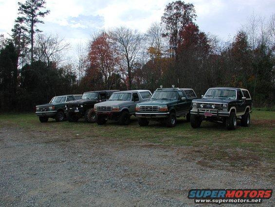 Several Ford Broncos and F-Series trucks parked in a grassy lot with trees in the background. The Bronco on the far right appears to be from the late 1980s