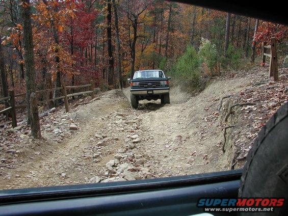Ford F-Series truck driving on a rocky trail in autumn woods. Another vehicle is partially visible in the foreground