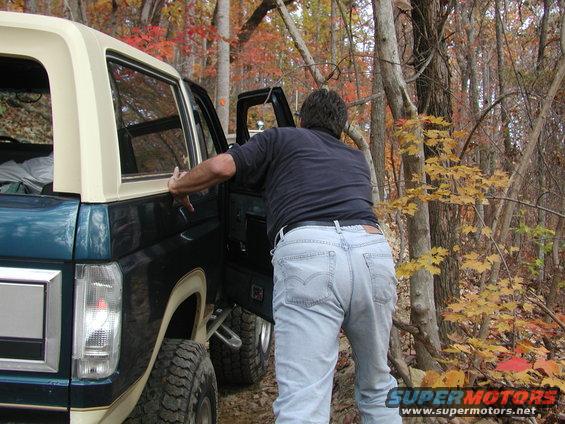 Dark teal and cream Ford Bronco with a man opening the driver's door, parked in a wooded area with fall foliage