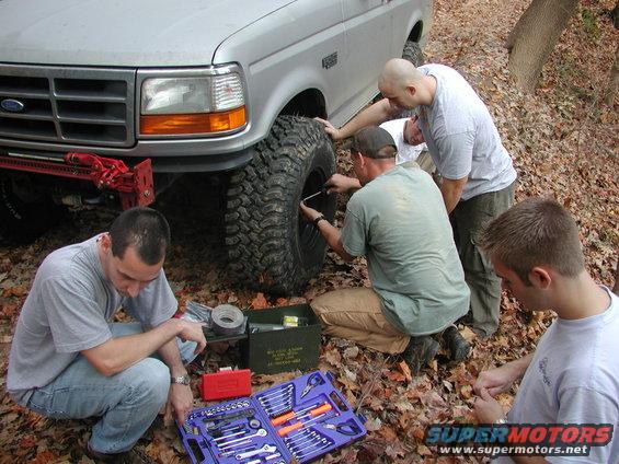 Silver truck front fender and tire with four men working on it. A red Hi-Lift jack is mounted on the front bumper