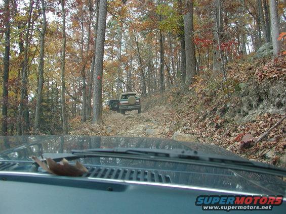 1985 Ford Bronco and another off-road vehicle driving on a rocky trail through autumn woods