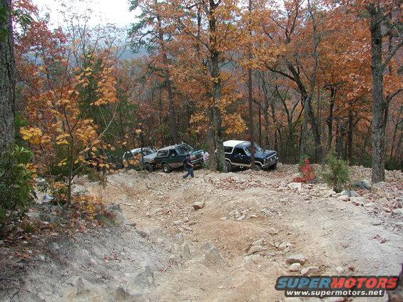 Several SUVs are driving on a rocky, wooded trail during autumn. One black Bronco is on the right, and a green Bronco and a white SUV are further back