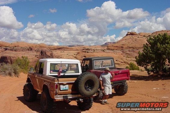 Two 1974 Ford Broncos parked on a dirt road in a desert with red rock formations. One is brown with a white top, the other is red