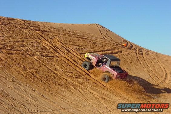 RC rock crawler with red and yellow body climbing a sandy hill, kicking up dust
