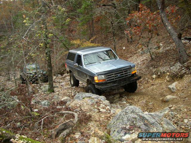 1995 Ford Bronco driving on a rocky, wooded trail. Another off-road vehicle is visible in the background
