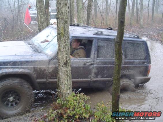 Muddy dark blue Jeep Cherokee XJ driving through water and mud, trees on the left, driver visible