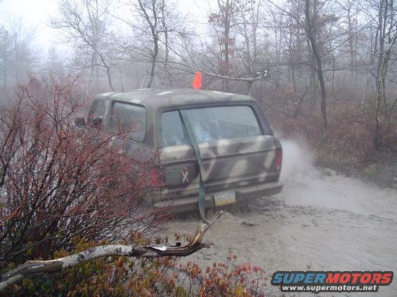 Camouflaged SUV stuck in mud on a dirt road, with trees and fog in the background