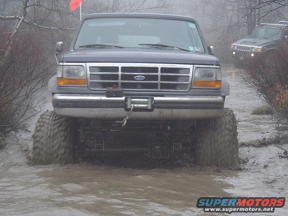 1993 Ford Bronco with large tires driving through a muddy trail, another SUV visible in the background