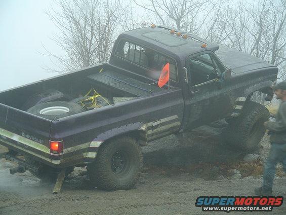 Purple pickup truck with lifted suspension stuck in mud, spare tire in bed. Text on rear says CHEVROLET