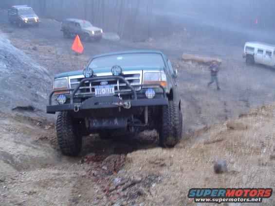 1993 Ford Bronco with aftermarket bumper and lights driving off-road, other SUVs and a white van in background
