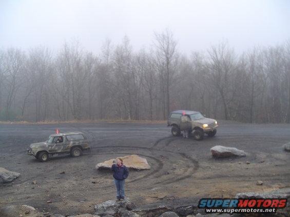 Two SUVs are parked on a dirt lot with trees in the background and fog. A person stands in the foreground
