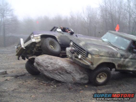 Two modified trucks are attempting to climb over a large boulder on a dirt course. One truck is purple, the other is camouflaged