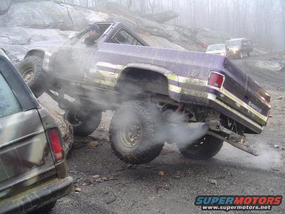 Two modified trucks off-roading in fog, one purple Chevrolet with large tires and exhaust smoke, next to a camouflage vehicle