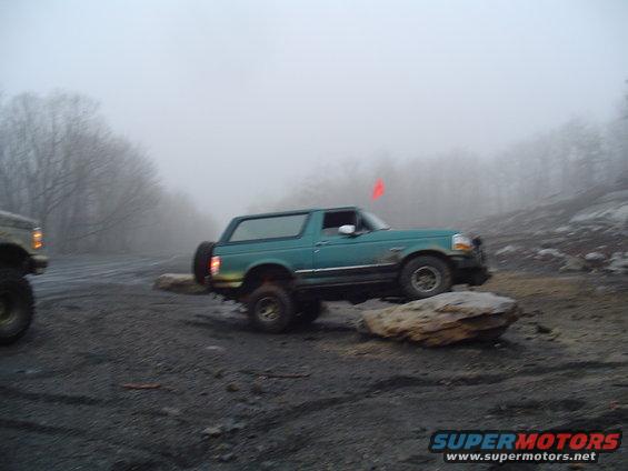 Teal 1993 Ford Bronco driving over rocks on a foggy day. Another vehicle is partially visible on the left