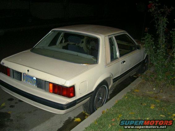 Rear view of a white 1982 Ford Mustang parked on the side of the road at night. The license plate is visible