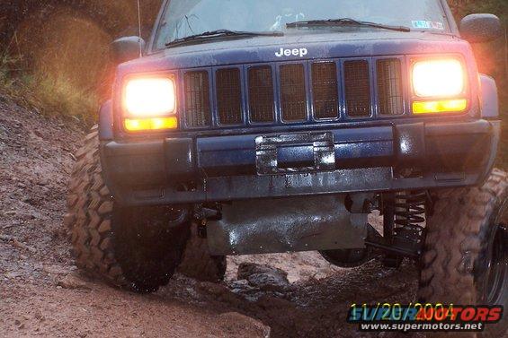 Blue Jeep Cherokee with headlights on, driving on a muddy trail