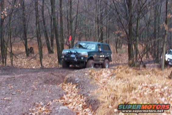 Green SUV with aftermarket bumper driving on a dirt trail in woods. Another white SUV is visible in the background