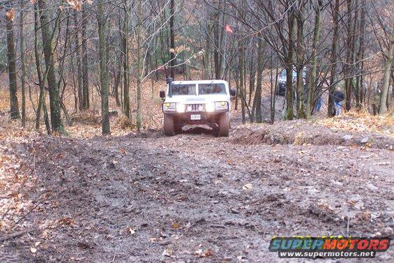 White SUV with snorkel and off-road tires navigates a muddy trail in a wooded area. Another vehicle is visible behind it
