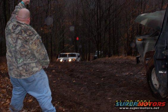 Guy in camo jacket watches a white Hummer H1 drive up a muddy hill in the woods. Another vehicle is partially visible on the right
