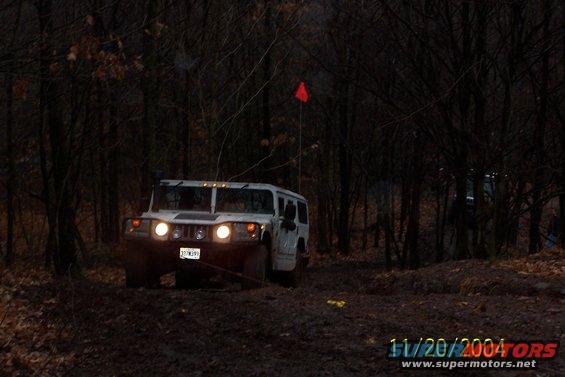 White SUV driving on a muddy trail in a dark forest, with headlights on and a red flag visible