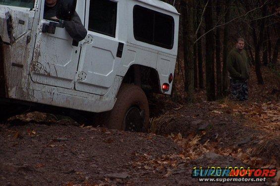 White Humvee stuck in mud on a wooded trail with a person standing in the background