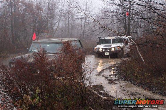 Two off-road vehicles driving through a muddy, wooded area on a foggy day