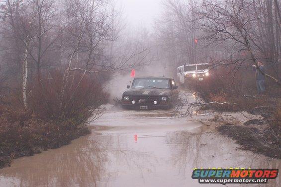 Dark 1978 Ford Bronco driving through muddy water, front view. Foggy conditions