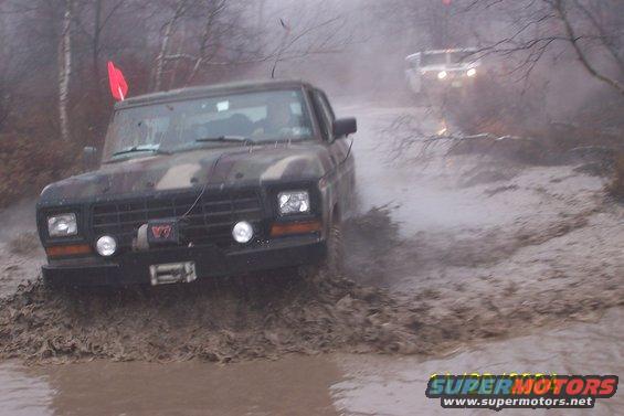 Camouflaged 1979 Ford Bronco driving through mud puddle, another vehicle in background