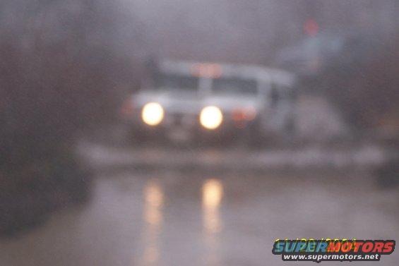 White SUV with headlights on driving through water, possibly in rain