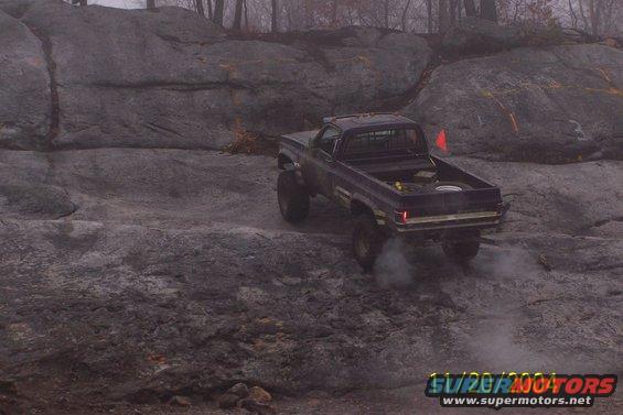 Dark blue pickup truck with lifted suspension driving over rocks, emitting smoke from rear tire