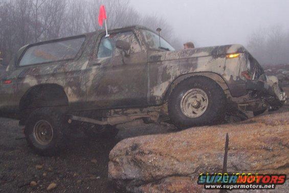 Mud-covered 1978 Ford Bronco on rocks, red flag on roof
