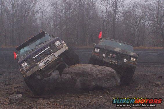 Two trucks are off-roading, one purple and one dark, navigating over a large rock. The purple truck has a chrome grille and bumper