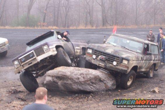 Two modified SUVs are attempting to drive over a large rock. One is purple and white, the other is camouflaged. A person is visible in the driver's seat of the purple vehicle