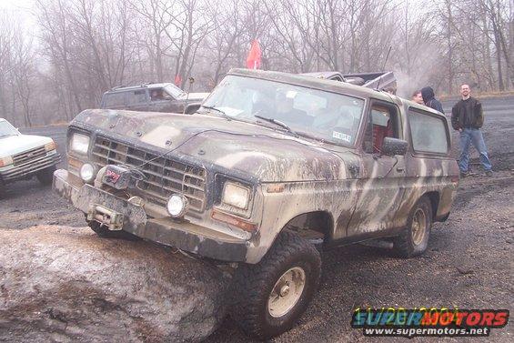 Muddy camouflage Ford Bronco climbing over a rock, another Bronco and a van in the background