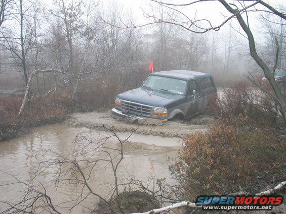 Dark blue Ford Bronco driving through a muddy water obstacle with trees and fog in the background