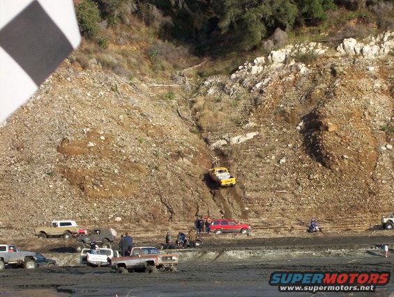 Several trucks and SUVs are stuck in mud at the bottom of a hill, with one yellow truck driving up the hill