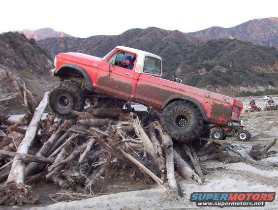 Red pickup truck with large tires climbing over a pile of logs, with ATVs in the background