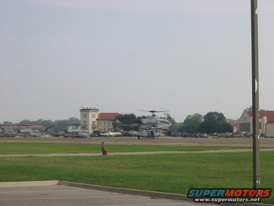 Military helicopters and fighter jets parked on an airfield with buildings in the background