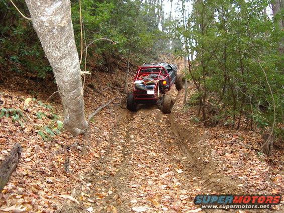 Off-road buggy with a red roll cage climbing a muddy, leaf-covered trail in a wooded area. Supermotors.net logo in lower right