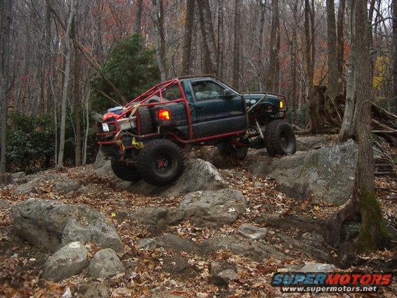 Modified off-road truck with a red roll cage climbing over rocks in a wooded area