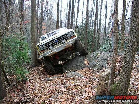Yellow Ford Bronco climbing over rocks on a wooded trail. A person stands on a rock ledge in the background