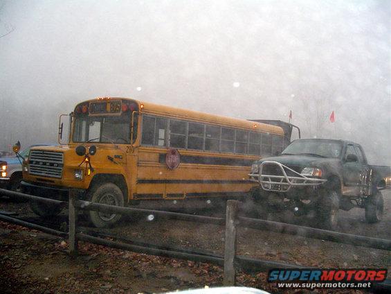 Yellow school bus parked next to a lifted pickup truck with a brush guard. Foggy conditions