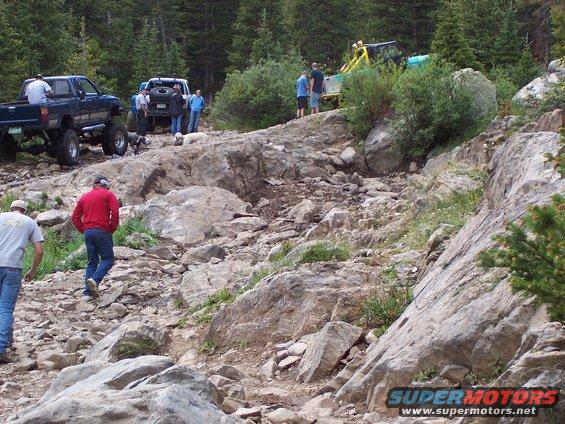 Several off-road vehicles navigate a steep, rocky mountain trail with people watching