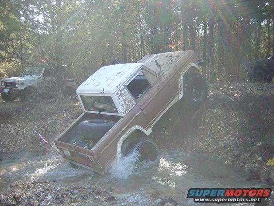 Brown and white 1974 Ford Bronco driving through water, with a spare tire in the bed