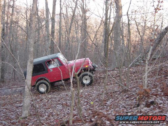 Red Jeep CJ-7 with black top driving on a wooded trail. Off-road tires and lifted suspension are visible