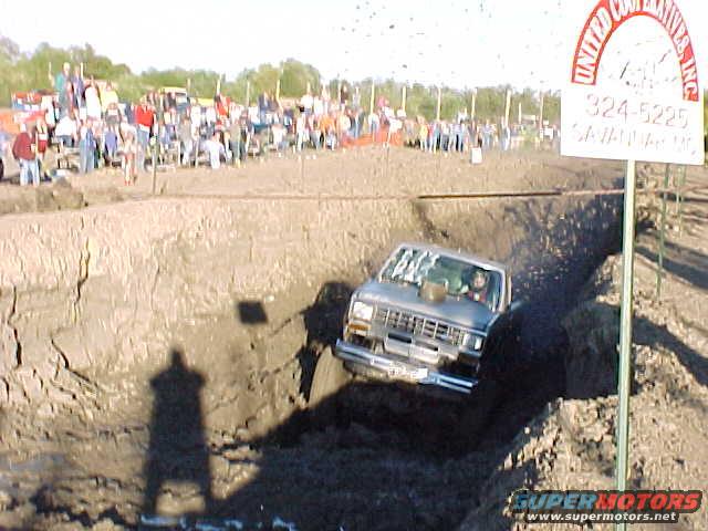 Truck navigating a mud pit with spectators in the background. Signage visible