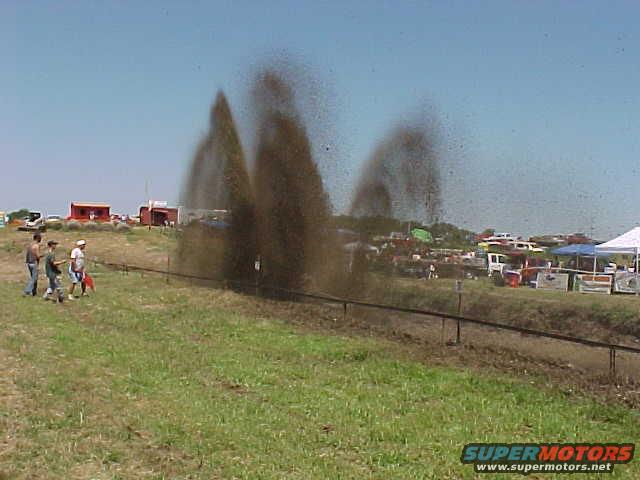 Off-road vehicles creating large mud spray at an event with spectators and tents in the background