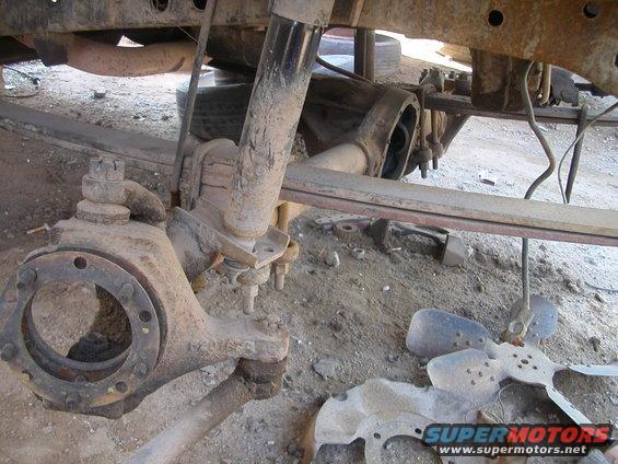 Close-up of a rusty front axle assembly with leaf springs and shock absorber. A fan blade lies on the ground