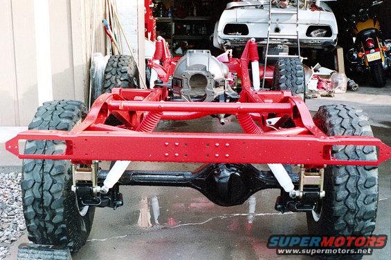 Red painted vehicle frame with large off-road tires, suspension components, and differential. Another vehicle body and a motorcycle are visible in the background