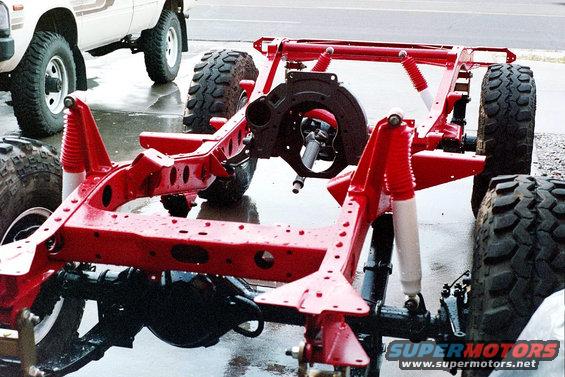 red painted vehicle frame with suspension components and tires, another vehicle in background